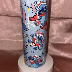 Puede incluir: Un vaso azul claro con un dise&ntilde;o festivo con el personaje Stitch con gorros y bufandas de Pap&aacute; Noel, copos de nieve y luces navide&ntilde;as. El vaso cil&iacute;ndrico est&aacute; sobre un pedestal blanco.