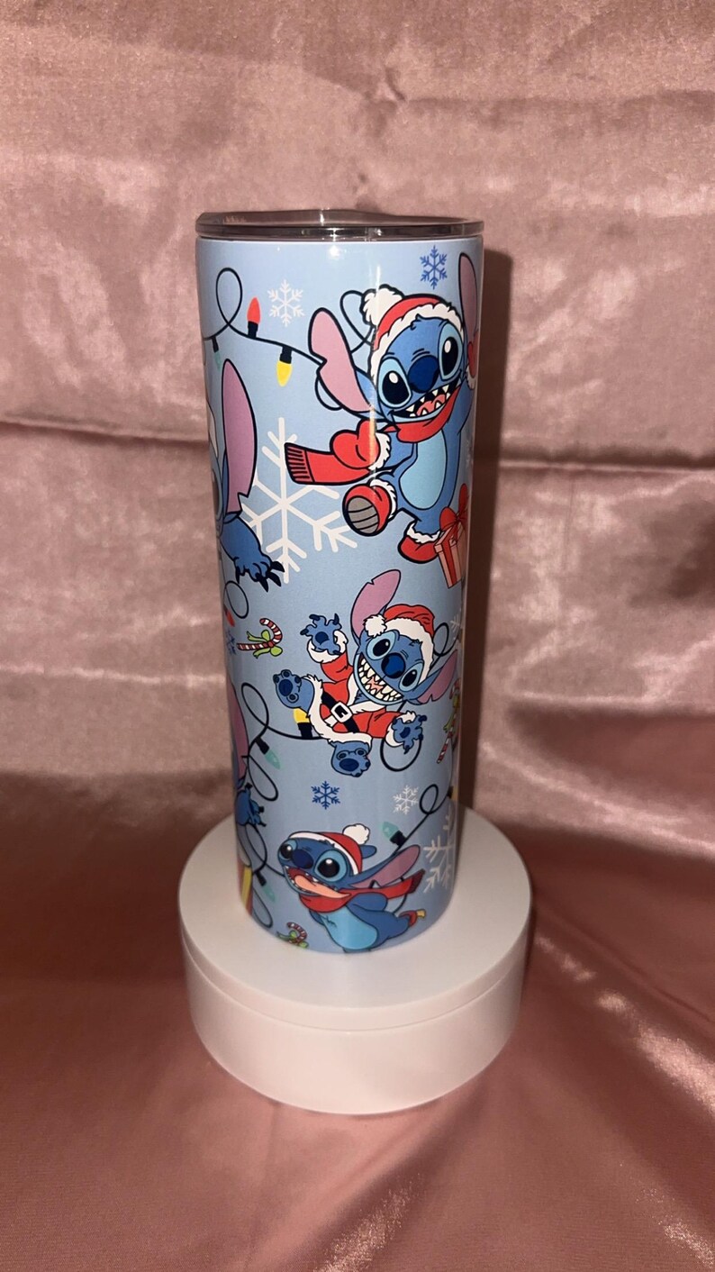 Puede incluir: Un vaso azul claro con un dise&ntilde;o festivo con el personaje Stitch con gorros y bufandas de Pap&aacute; Noel, copos de nieve y luces navide&ntilde;as. El vaso cil&iacute;ndrico est&aacute; sobre un pedestal blanco.
