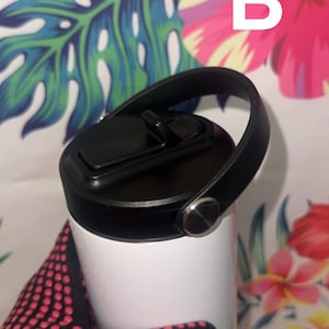 Puede incluir: Un vaso blanco con tapa y asa negras. La tapa tiene una abertura abatible. El asa est&aacute; sujeta con un bot&oacute;n plateado. La letra "B" es visible en blanco. El fondo presenta un estampado floral.