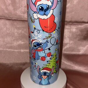 Puede incluir: Un vaso azul claro con el personaje Stitch de Lilo & Stitch con gorros de Pap&aacute; Noel y decoraciones navide&ntilde;as. El vaso tiene una tapa plateada y se muestra sobre un soporte blanco.
