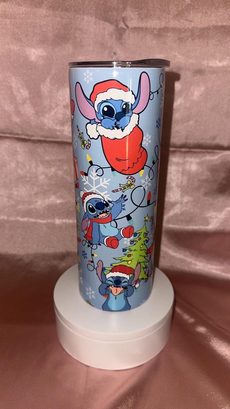 Puede incluir: Un vaso azul claro con el personaje Stitch de Lilo & Stitch con gorros de Pap&aacute; Noel y decoraciones navide&ntilde;as. El vaso tiene una tapa plateada y se muestra sobre un soporte blanco.