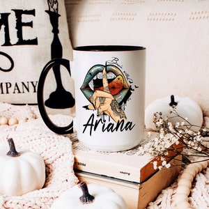 May include: White ceramic mug with a black handle and rim. The mug features a colorful illustration of a pair of lips with fangs and a hand with a finger over the lips. The text "Basic witch" is above the lips and the name "Ariana" is below the lips.