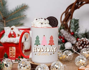 May include: A white ceramic mug with a red interior and handle, decorated with Christmas trees and the words "HOLIDAY cheer". The mug is filled with whipped cream, chocolate chips, and a cookie. Decorative disco ball lights and a wreath are in the background.