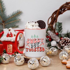 May include: A white ceramic mug with a red handle and rim, filled with whipped cream and a cookie. The mug features the text "Sweet But Twisted" in pink, black, and red. The mug is on a wooden surface with decorative lights and ornaments.