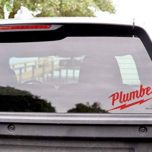 May include: A red "Plumber" decal with a lightning bolt design is affixed to the rear window of a white truck. The decal is visible against the backdrop of the vehicle's interior and the outside environment.