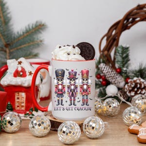 May include: A large white mug with a red handle and rim, featuring three nutcracker skeleton figures in red, purple, and black uniforms. The mug is filled with whipped cream and an Oreo cookie. The text "LET'S GET CRACKIN'" is below the figures.