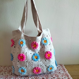 May include: A crocheted tote bag with a light beige background and colourful flower designs. The bag has two straps and is made with a granny square pattern.