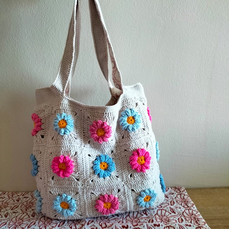 May include: A crocheted tote bag with a light beige background and colourful flower designs. The bag has two straps and is made with a granny square pattern.