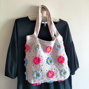 May include: A crocheted tote bag with a floral design. The bag is made of beige yarn and features pink, blue, and white flowers.