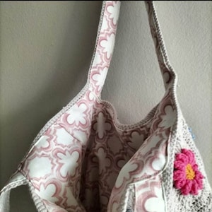 May include: A crocheted tote bag with a pink floral lining and a pink crocheted flower accent. The bag has a long strap and is made of a beige yarn.