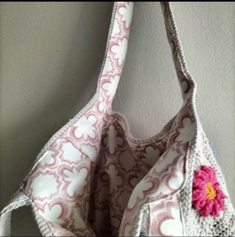 May include: A crocheted tote bag with a pink floral lining and a pink crocheted flower accent. The bag has a long strap and is made of a beige yarn.