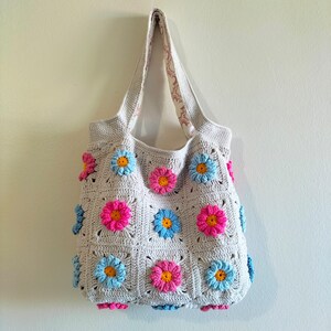 May include: A crocheted tote bag with a light grey background and colourful flower designs. The bag has a light pink, light blue, and yellow flower pattern.