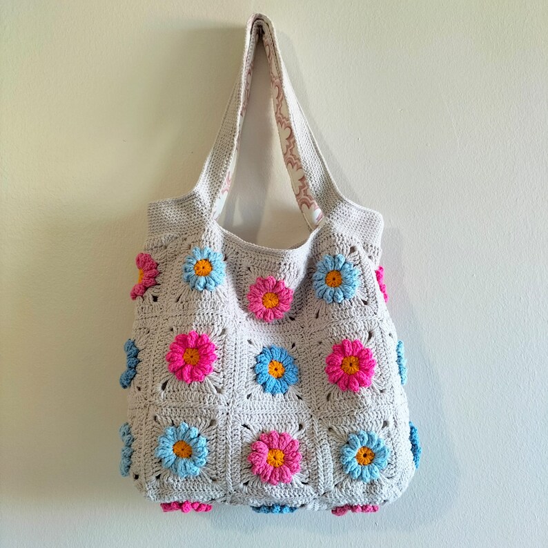 May include: A crocheted tote bag with a light grey background and colourful flower designs. The bag has a light pink, light blue, and yellow flower pattern.