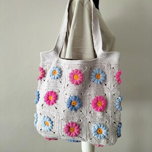 May include: A crocheted tote bag with a light grey background and colourful flower designs. The bag has two long straps and is made with a granny square pattern.