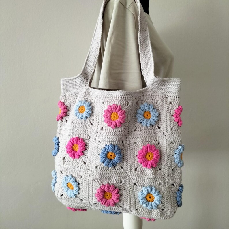 May include: A crocheted tote bag with a light grey background and colourful flower designs. The bag has two long straps and is made with a granny square pattern.