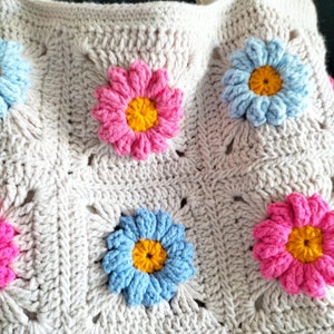 May include: A crocheted bag with a beige background and colourful flower accents. The flowers are pink, blue, and yellow.