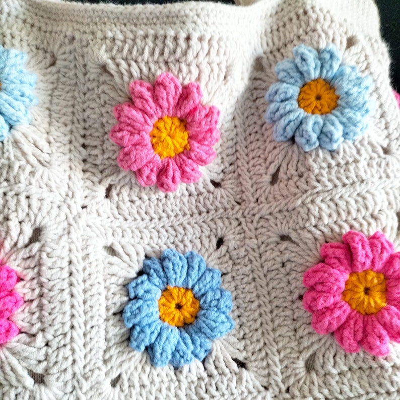 May include: A crocheted bag with a beige background and colourful flower accents. The flowers are pink, blue, and yellow.