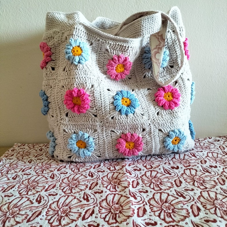 May include: A crocheted tote bag with a light grey background and colourful flower designs. The flowers are pink, blue and yellow.