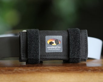 Könnte beinhalten: Eine schwarze Halsbandhülle aus Stoff mit zwei vertikalen Klettverschlussriemen. Ein quadratisches Etikett ist vorne mittig angebracht und zeigt eine sonnenuntergangsfarbene ovale Grafik mit der Silhouette eines Hundes und dem Text Unconditional Love animal bodywear.