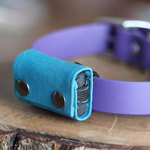 May include: A teal fabric dog waste bag holder attached to a purple leather dog collar. The holder has two metal snap closures. The collar has a silver metal buckle and hardware. The items are on a wooden surface.