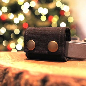May include: A close-up of a black canvas belt loop with two brass-colored snap closures. A gray belt is partially visible, extending from the loop. The background features a blurred Christmas tree with colorful lights, set on a wooden surface.