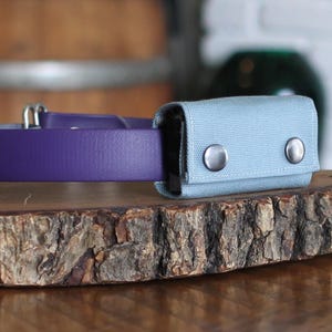 May include: A purple collar made of a smooth, synthetic material featuring a light blue rectangular fabric pouch attached to the side. The pouch is secured with two silver-toned metal snaps on the front flap and is mounted onto the collar strap.
