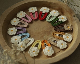 May include: A set of 15 crocheted flower hair clips in various colors, arranged in a circle on a wooden surface. Each clip features a white crocheted flower with a yellow center.