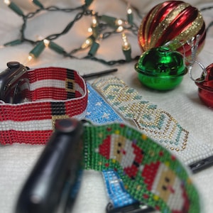 May include: A black smartwatch with a red and white Santa Claus-themed band, alongside other beaded watch bands with festive designs. Christmas ornaments and string lights are in the background, creating a holiday theme.