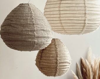 May include: Three hanging pendant lights with a textured, woven fabric design. The lampshades are in varying sizes and shades of beige and tan. The largest lamp is a light beige, while the others are a darker tan color. The lamps are suspended from the ceiling.