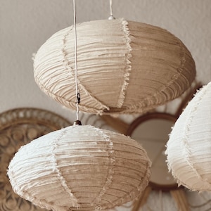 May include: Three beige fabric pendant lights hanging from the ceiling. The lampshades have a round, oval shape with a textured, fringed edge. The background includes decorative wall hangings and a mirror.