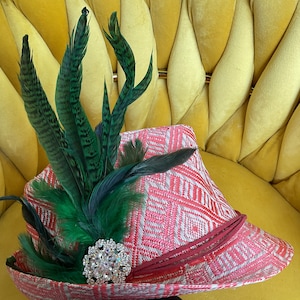 Könnte beinhalten: Hut mit rosa-weißem Muster, verziert mit grünen und schwarzen Federn und einer Strassbrosche. Der Hut hat ein rotes Band. Ein stilvolles Modeaccessoire.