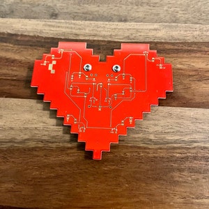 May include: A red heart-shaped circuit board with gold-colored traces and two silver screws. The board has a pixelated edge.