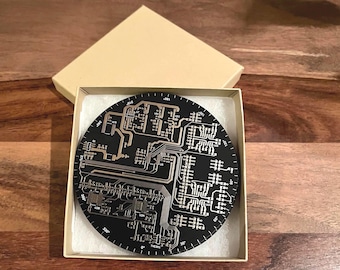 May include: A round, black coaster with a silver circuit board design. The coaster is in a beige box with white tissue paper.