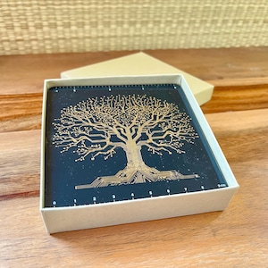 May include: Square coaster with a gold tree design on a black background, presented in a beige box. The tree's branches and roots are intricately detailed. The coaster measures 9 cm.