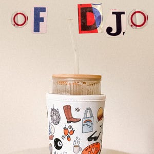 以下が含まれることがあります： 木製の蓋と白い布製のスリーブが付いたガラスタンブラー。スリーブは、ディスコボール、カウボーイブーツ、レインボーのトートバッグ、その他の楽しいデザインのカラフルなイラストで装飾されています。「CUP OF DJO」の文字がタンブラーの上に表示されています。