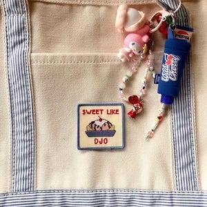 May include: A beige canvas tote bag with blue and white striped trim, featuring a square embroidered patch that says SWEET LIKE DJO above a banana split illustration. A beaded keychain with a pink character and a blue Wild Cherry Pepsi tube.