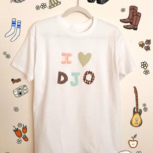 May include: White t-shirt with the text "I ♥ DJO" in colourful, patchwork-style letters. The shirt is hanging on a hanger, surrounded by illustrated doodles of a disco ball, sunglasses, boots, and other objects.
