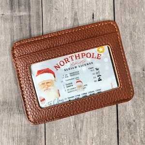 May include: A brown leather wallet with a clear plastic window displaying a fake North Pole Sleigh License. The license is for Claus Santa, with an expiration date of 05/08/2026, and an address of 25 Candy Cane Lane, North Pole 33907.