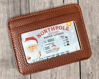 May include: A brown leather wallet with a clear plastic window displaying a fake North Pole Sleigh License. The license is for Claus Santa, with an expiration date of 05/08/2026, and an address of 25 Candy Cane Lane, North Pole 33907.