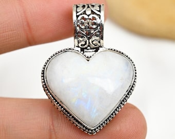 Puede incluir: Un colgante de plata en forma de corazón con una piedra de luna blanca. El colgante tiene un diseño filigrana intrincado alrededor de la piedra.