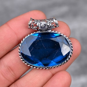 Może przedstawiać: Srebrny wisiorek z dużym, owalnym, fasetowanym niebieskim kamieniem szlachetnym osadzonym w srebrnej oprawie. Wisiorek ma delikatny kwiatowy wzór wokół kółka.