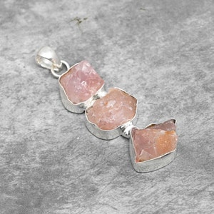 Può includere: Un ciondolo d'argento con tre pietre di quarzo rosa grezzo. Le pietre sono di colore rosa chiaro e sono incastonate in argento.