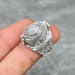 May include: A silver-colored ring with an oval-shaped, light-colored stone at the center. The ring features intricate, ornate detailing and a hinged top. The ring is shown on a finger against a gray background.