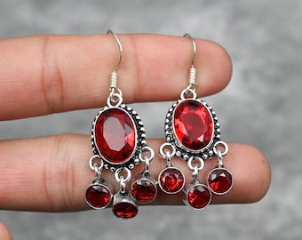 Pode incluir: Um par de brincos de prata pendurados com detalhes de pedras preciosas vermelhas. Os brincos apresentam uma pedra preciosa vermelha de forma oval grande cercada por pedras preciosas vermelhas menores.