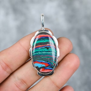 May include: A silver pendant featuring an oval-shaped, multi-coloured stone. The stone has a layered pattern of black, green, blue, red, and white.