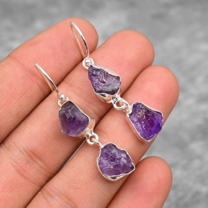 May include: A pair of silver dangle earrings with two raw amethyst stones on each earring. The amethyst stones are a deep purple color.