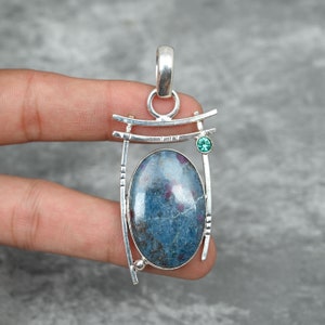 以下が含まれることがあります： 銀製のペンダントに、青と白の楕円形の宝石が銀の枠にセットされています。枠の上部右側に小さな緑色の宝石が付いています。