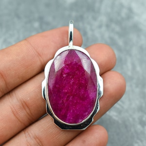 May include: A silver pendant featuring a large, oval-shaped ruby gemstone. The ruby is a deep, rich red colour and has a faceted cut. The pendant is set in a silver frame with a scalloped edge.