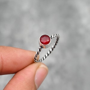 Può includere: Un anello d'argento con una gemma rotonda di colore rosso vivo. L'anello ha un design a fascia intrecciata. La gemma è incastonata in una lunetta d'argento. L'anello è tenuto da un dito.