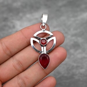 Pu&ograve; includere: Un ciondolo d'argento con un design geometrico che presenta due pietre preziose rosse. Il ciondolo ha la forma di un fiore stilizzato con una pietra preziosa a forma di lacrima che pende dal centro.